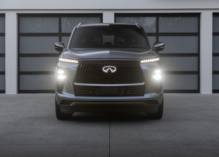 A front view of the 2025 INFINITI QX80 parked in front of multiple windows with the headlights and emblem lit.