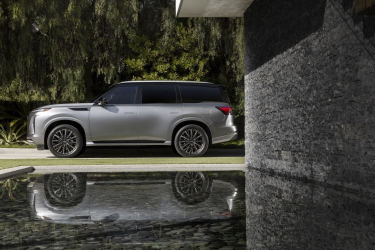 A left side view of a silver 2025 INFINITI QX80 parked next to water and greenery, its reflection can be seen in the water.